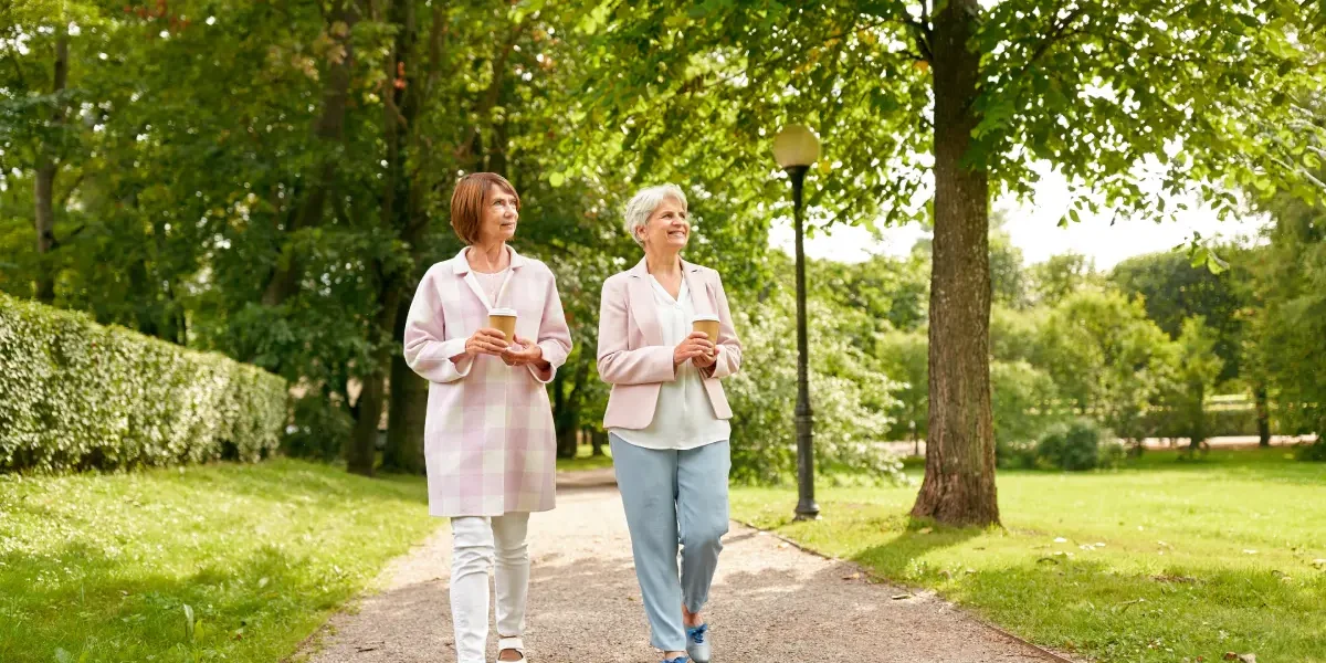 Two senior women walking through a park holding drinks
